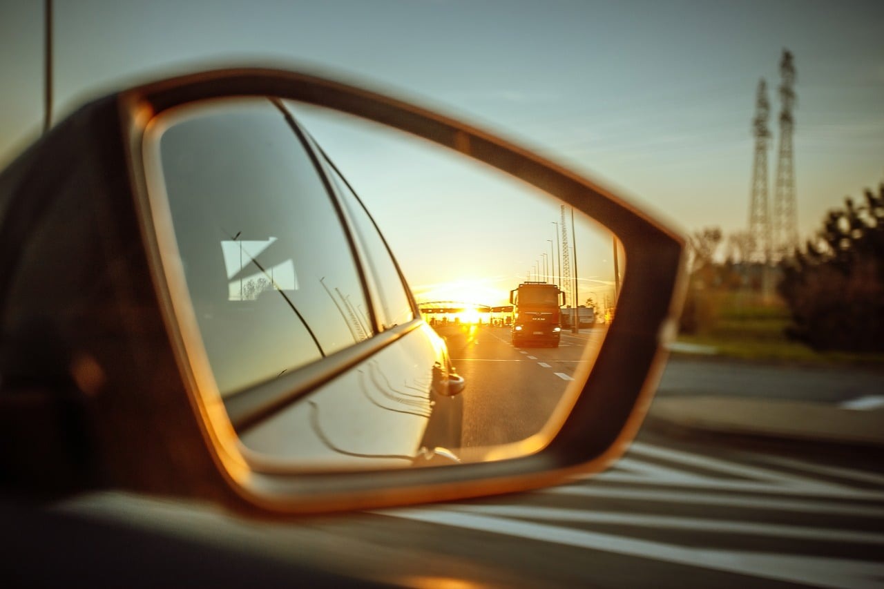 A car side mirror frames sunset traffic, utility lines, and the bright edge of a white vehicle beside a blurred roadway.
