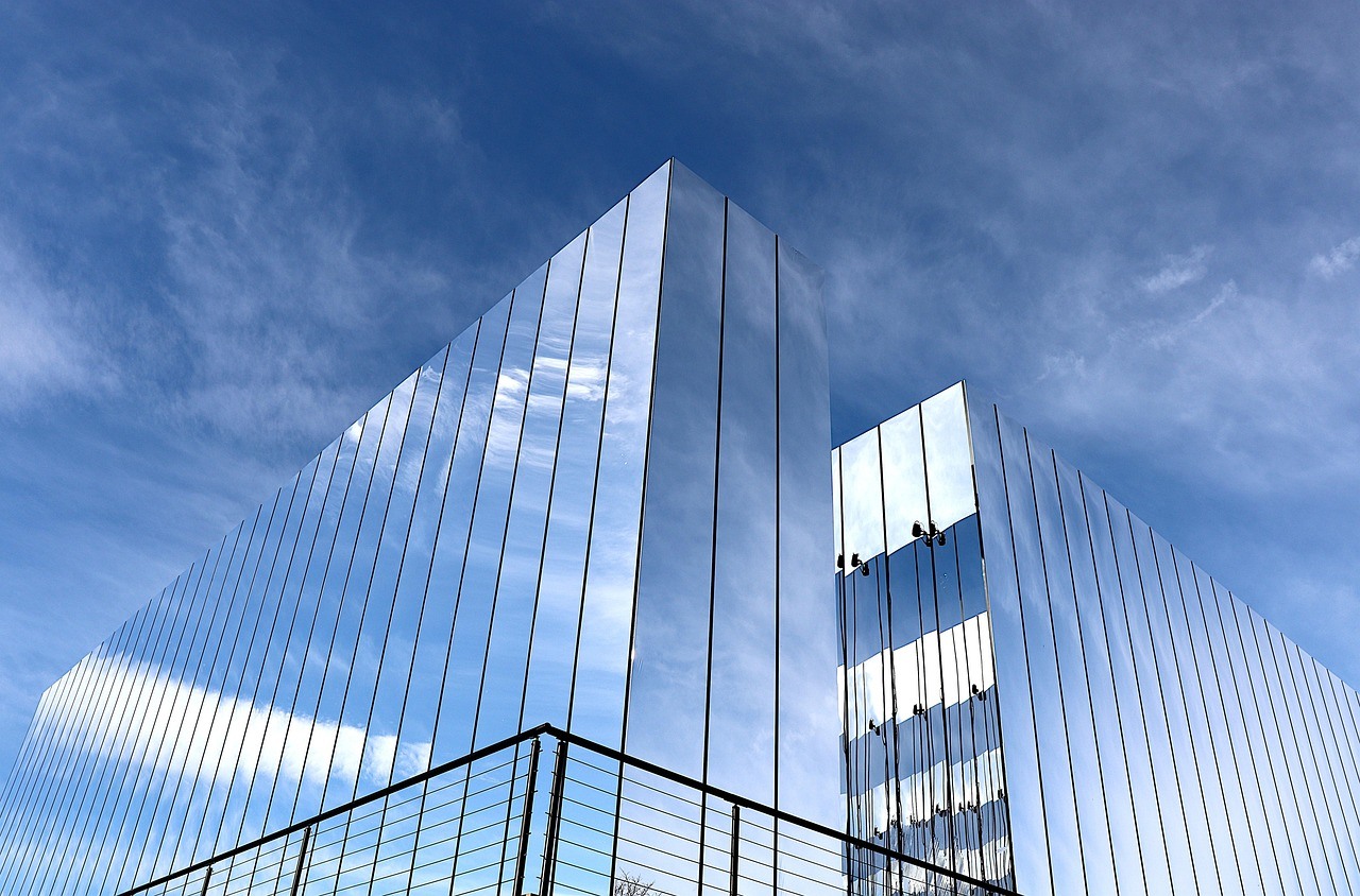 A modern glass building rises beneath blue clouds, its reflective vertical panels dividing sky and light into narrow sections.